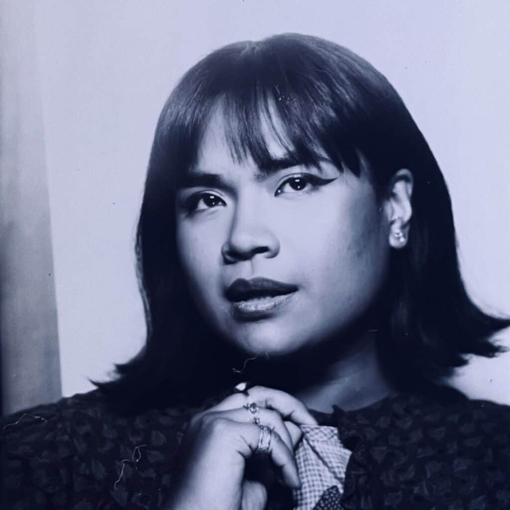 A black-and-white headshot of Denny, an Asian woman in her 20s. She sports winged eyeliner and a bob, and holds her ring-laden hand close to her chin.