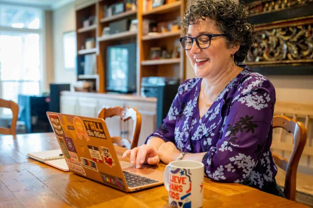 Mandy, a white woman with short, dark curly hair and black-rimmed glasses, types on her. laptop in her home. Her laptop is covered in LGBTQ+ pride stickers, and next to her is a mug that reads "Parents of Trans Youth."