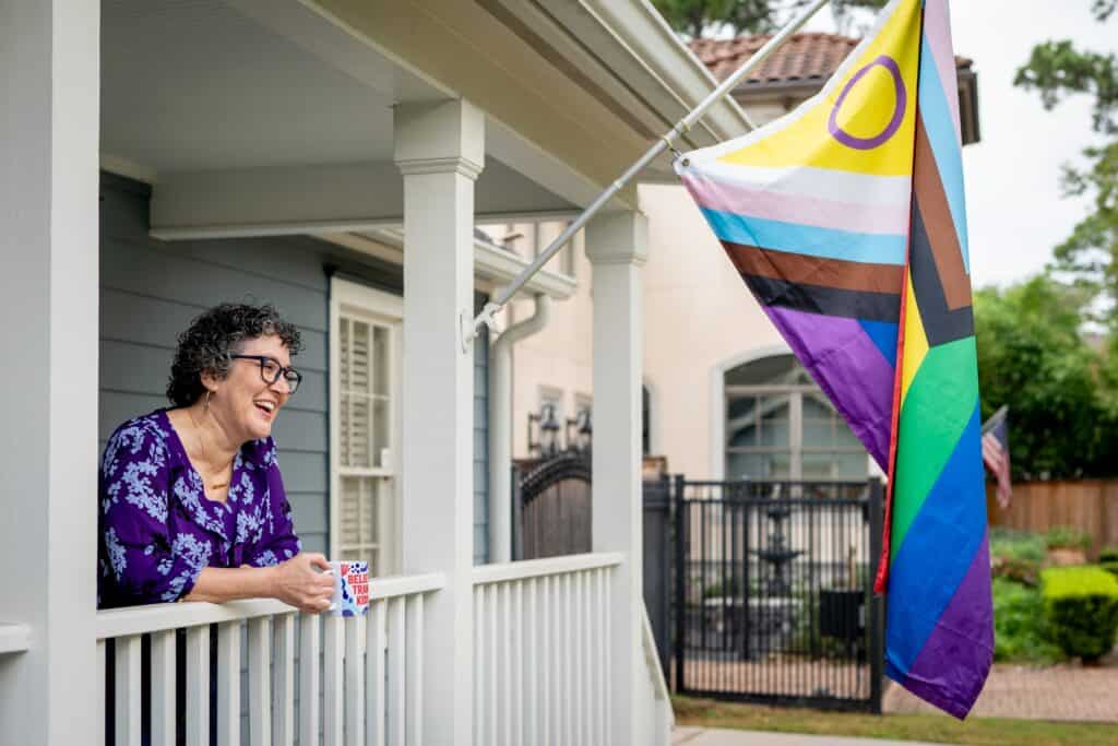 Mandy Giles, a white woman with dark curly hair and glasses, stands on her white-fenced veranda, looking out at the yard. She has an open-mouthed smile as though talking to someone, and she is holding a mug that reads, "Believe Trans Kids." Next to her flies an LGBTQI+ flag with rainbow, black, brown, white, pastel blue, and pastel pink stripes, and a yellow triangle with a purple circle in it.