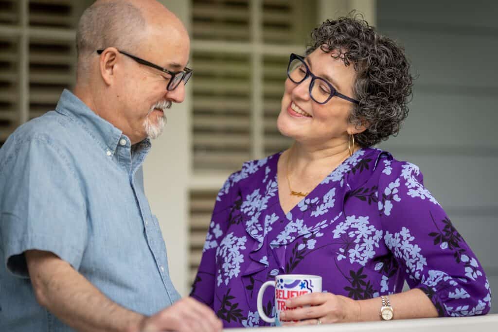 Mandy and Neil Giles lovingly look at each other on the veranda of their home. Both wear dark-rimmed glasses. Neil wears a light blue short-sleeved button-up, and Mandy wears a purple patterned blouse.