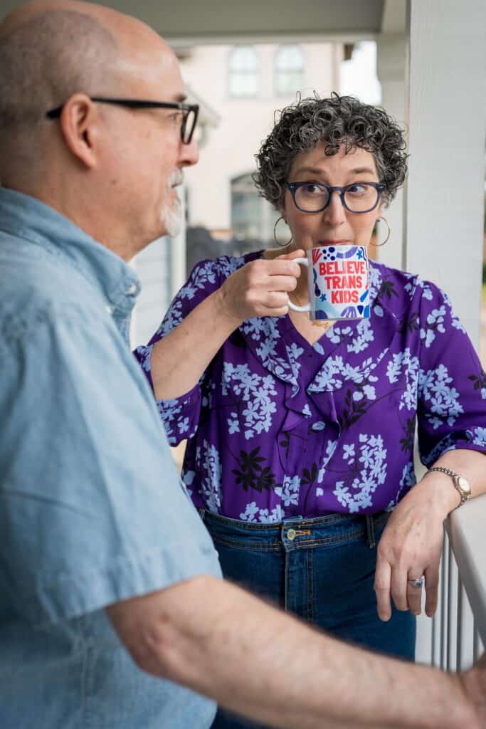 Mandy Giles, a white woman with dark curly hair, looks at her husband, Neil, a bald white man with a gray beard, on the veranda of their home. Mandy has an amused expression on her face as she sips from a mug that reads, “Believe Trans Kids.” Neil is wearing a light blue short-sleeved button-up, and Mandy wears a purple patterned blouse. Both are wearing dark-rimmed glasses.