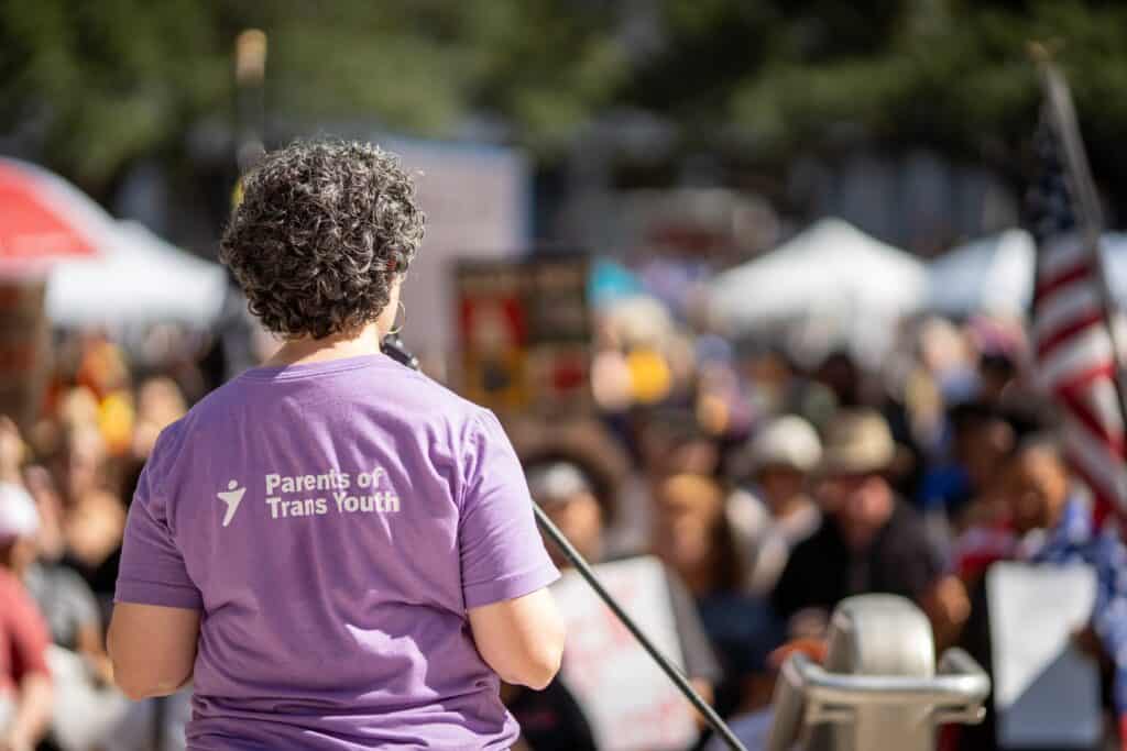 Mandy Giles, a white woman with short, dark curly hair, stands in front of a microphone and crowd at a public demonstration. She is facing away from the camera, and the back of her lavender shirt reads, "Parents of Trans Youth"