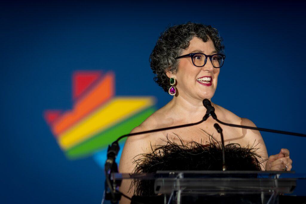 Mandy, a white woman with dark curly hair, stands at a lectern and microphone. Behind her is the Equality Texas Logo, which is the shape of Texas with rainbow stripes. Mandy is wearing a strapless black feathered top.