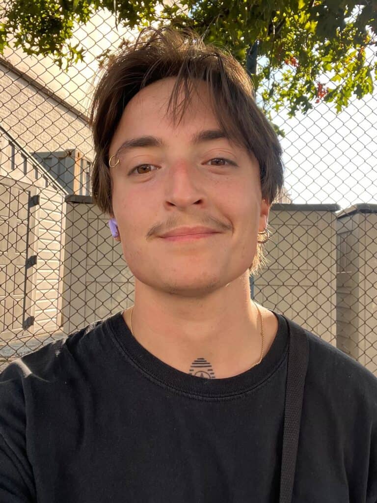 A young, white transmasculine person stands in front of a chain link fence. They have short brown hair, a gold hoop eyebrow ring, and a small tattoo poking above the collar of their black T-shirt.