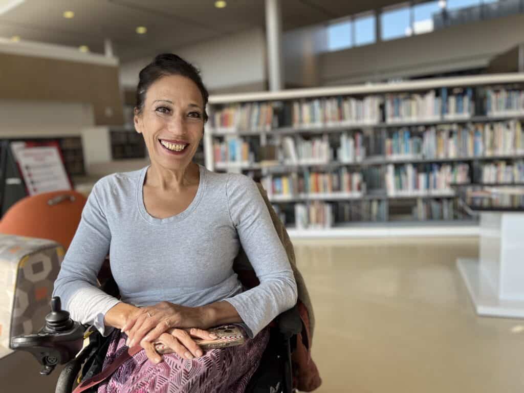 Maria, a 59-year-old Latina woman, is posed in front of a white shelf of books at a library. She is sitting in a motorized wheelchair with her hands crossed in her lap, and she is wearing a long-sleeved gray shirt. Her mauve print pants match her lipstick. Her hair is pulled back, and she is smiling widely. 