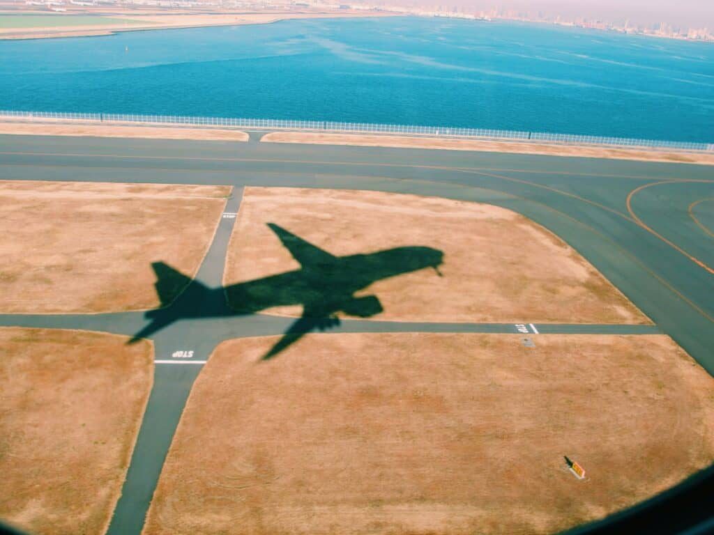 The shadow of an airplane taking off on a clear day, over a runway by a bright blue sea.