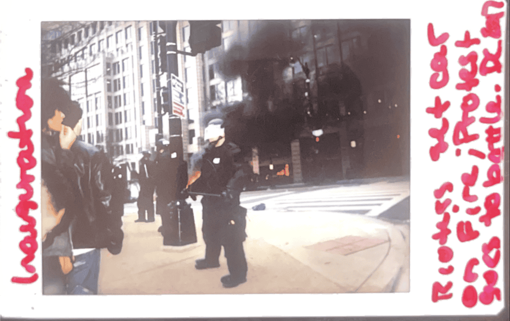 A horizontal Polaroid of a police officer in riot gear, standing on a city street corner. Behind the officer is a building clouded by black smoke from a car that has been set on fire. Red writing on the edges of the Polaroid reads: Inauguration. Rioters set car on fire; protest goes to battle. D.C. 2017