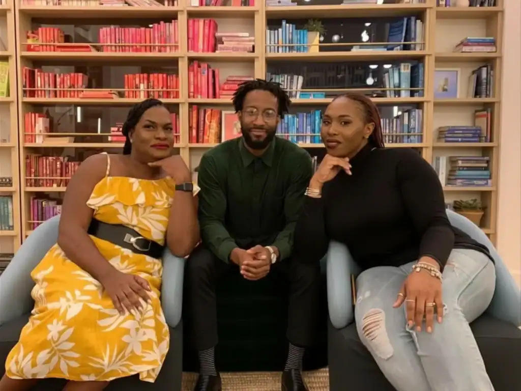 TransLash founder Imara Jones, a black woman in a floral summery dress, activist Nala Simone, black woman in a turtle neck and Justin Freeman, a black man in glasses, sitting in front of a bookshelf, posing together for the camera